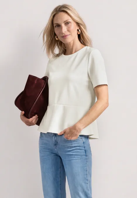 Shirt met ronde hals en volantdetail off white Vrouw in wit peplumshirt met korte mouwen en spijkerbroek, houdt een bordeaux clutch vast.
