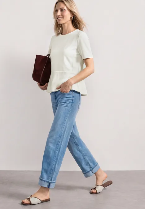 Shirt met ronde hals en volantdetail off white Vrouw met witte peplumtop, lichtblauwe opgerolde jeans, platte sandalen en een bordeaux clutch.
