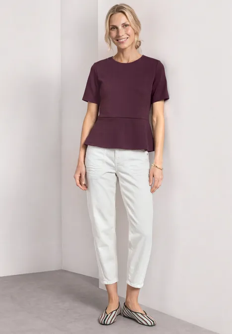 Shirt met ronde hals en volantdetail rustic rouge Vrouw in paarse top, witte broek en gestreepte ballerina's tegen neutrale achtergrond.