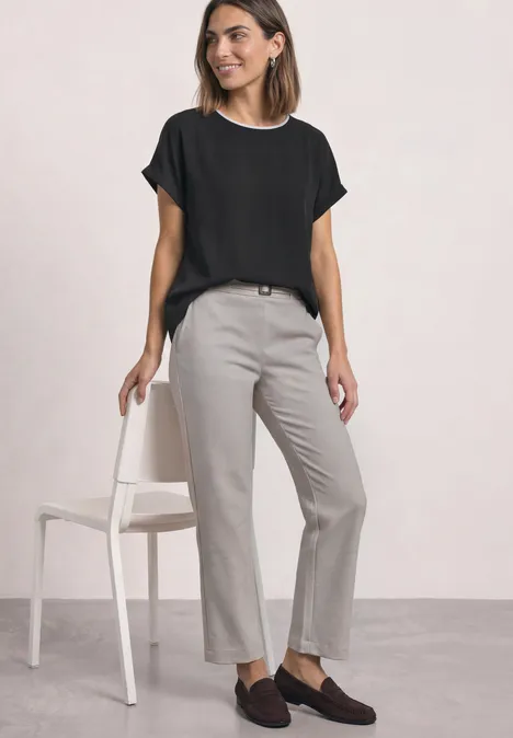 2-in-1 Shirt aus Chiffon mit Boat Neck Black Frau trägt schwarze Bluse, graue Hose und braune Loafers, steht lässig neben Stuhl.