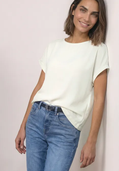 2-in-1 Shirt aus Chiffon mit Boat Neck off white Frau steht vor hellem Hintergrund, trägt weißes T-Shirt und blaue Jeans, lächelt entspannt.
