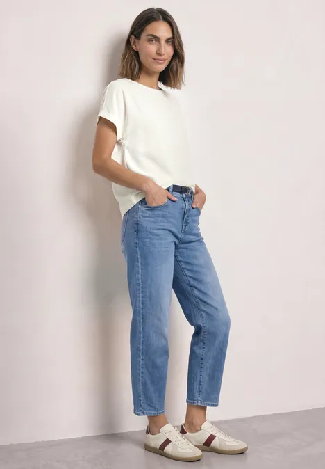 2-in-1 Shirt aus Chiffon mit Boat Neck off white Eine Frau trägt ein weißes T-Shirt, blaue Jeans und weiße Sneakers und posiert vor hellem Hintergrund.