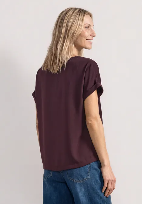 Rückansicht einer Frau in lila Bluse und Jeans, lächelnd, den Kopf zur Seite gedreht.
