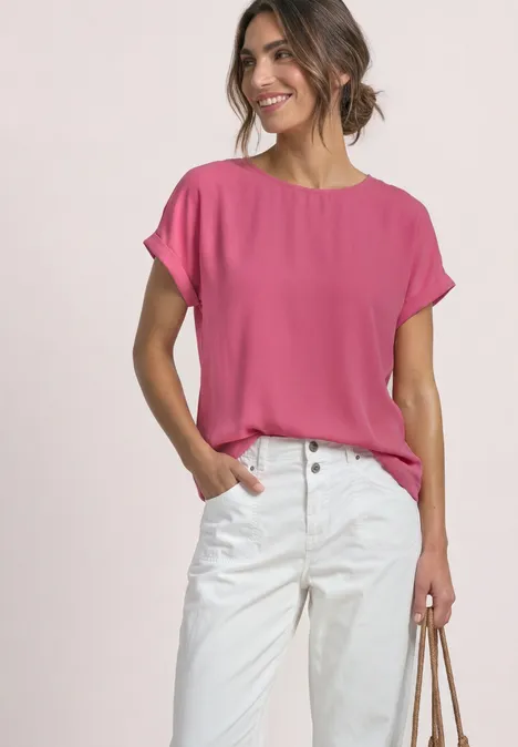 Frau trägt pinkes kurzärmeliges Oberteil und weiße Hose, lächelt, Hand in Tasche, lässiger Look.