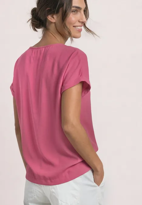 2-in-1 Shirt aus Chiffon mit Boat Neck blush berry Eine seitliche Aufnahme einer lächelnden Frau in pinkem T-Shirt und weißen Hosen mit Händen in den Taschen.