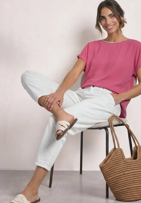 2-in-1 Shirt aus Chiffon mit Boat Neck blush berry Eine Frau sitzt entspannt auf einem Stuhl in weißer Hose, pinkem Shirt und Sandalen mit Korbtasche.