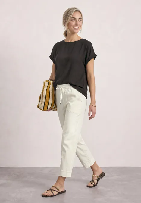 2-in-1 Shirt aus Chiffon mit Boat Neck black coffee Frau in schwarzem Shirt, hellem Bundfaltenhose und Sandalen mit gestreifter Tasche, lächelnd spazierend.
