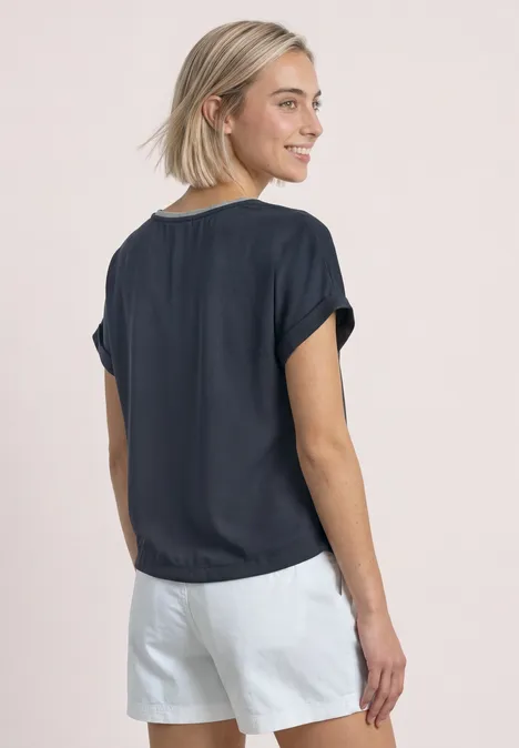 Frau mit kurzem blonden Haar trägt legeres dunkelblaues Shirt und weiße Shorts, seitlich von hinten.