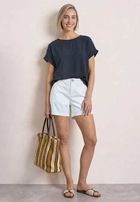 Eine lächelnde Frau in dunkelblauem Shirt, weißen Shorts und Sandalen trägt eine gestreifte Tasche.