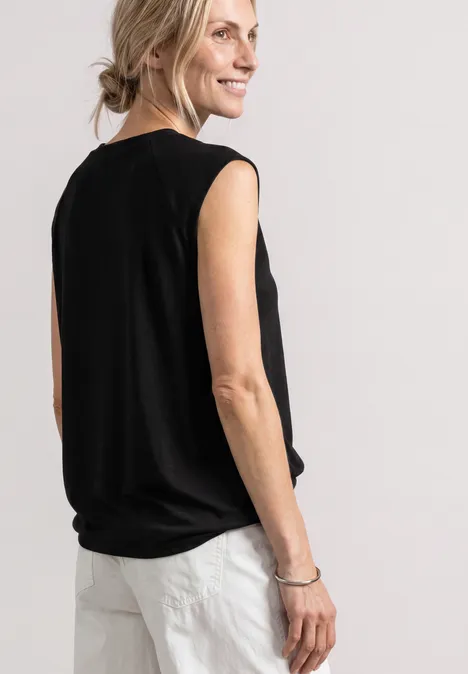 Een vrouw in zwart mouwloos topje en witte broek kijkt glimlachend over haar schouder.