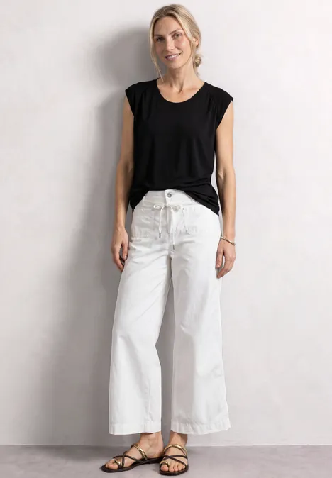 Vrouw in een zwart mouwloos topje, witte wijde broek en sandalen, glimlachend voor een neutrale achtergrond.
