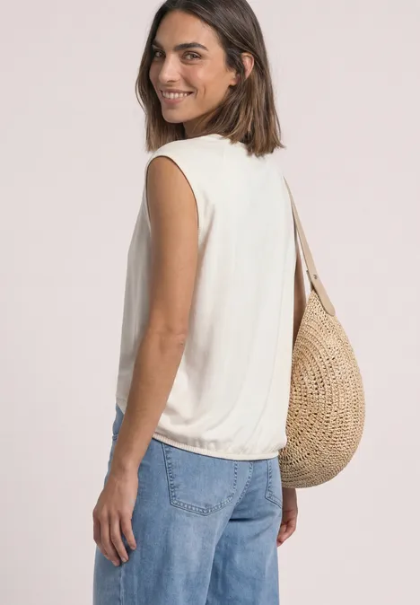 Frau in weißem ärmellosem Top und Jeans, mit Strohtasche, seitlich lachend abgelichtet.