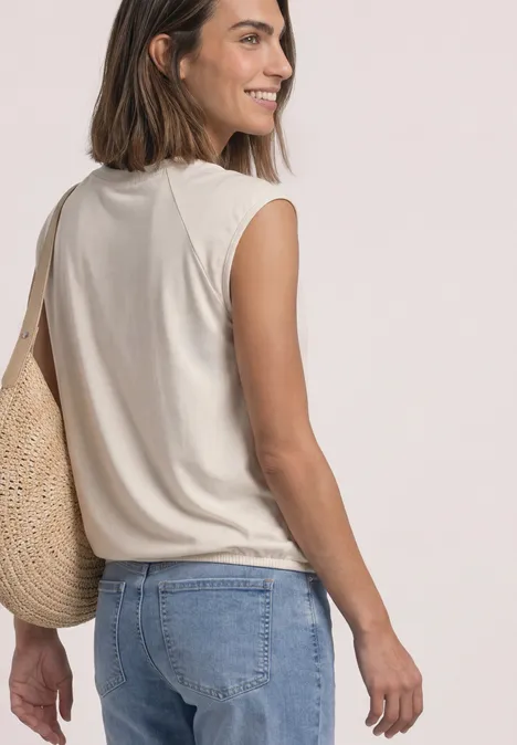 Frau in ärmellosem Top, Jeans und Strohtasche lächelt über die Schulter vor hellem Hintergrund.
