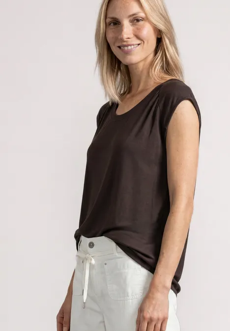 Glimlachende vrouw met bruin topje en witte broek voor een lichte achtergrond.