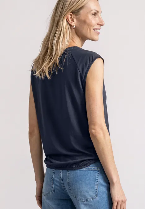 Une femme blonde en haut sans manches noir et jean bleu vue de profil, souriante, regardant vers la droite.
