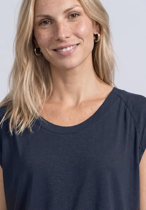 Femme blonde souriante avec taches de rousseur en haut bleu foncé sur fond gris clair.