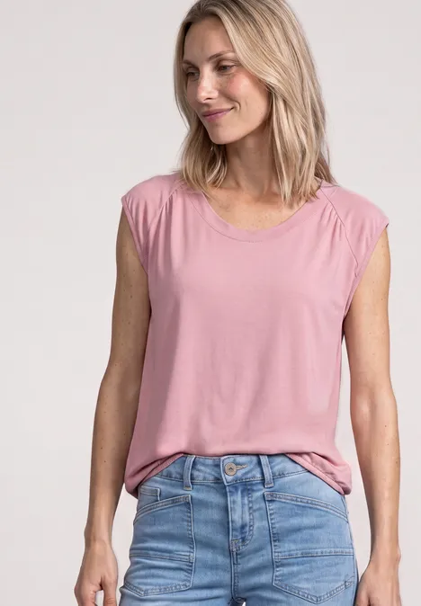 Blondine lacht in een mouwloze roze top en een blauwe spijkerbroek.