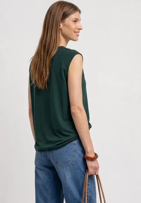 Een vrouw draagt een donkergroen mouwloos topje en een blauwe jeans, houdt een tas vast en kijkt zijwaarts.