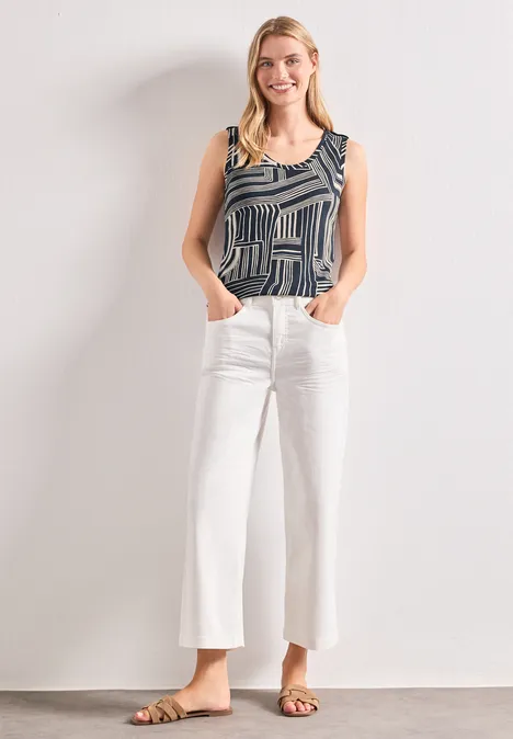 Frau in gemustertem Top, weißen weiten Cropped-Jeans und Sandalen steht mit Händen in den Taschen vor weißer Wand.
