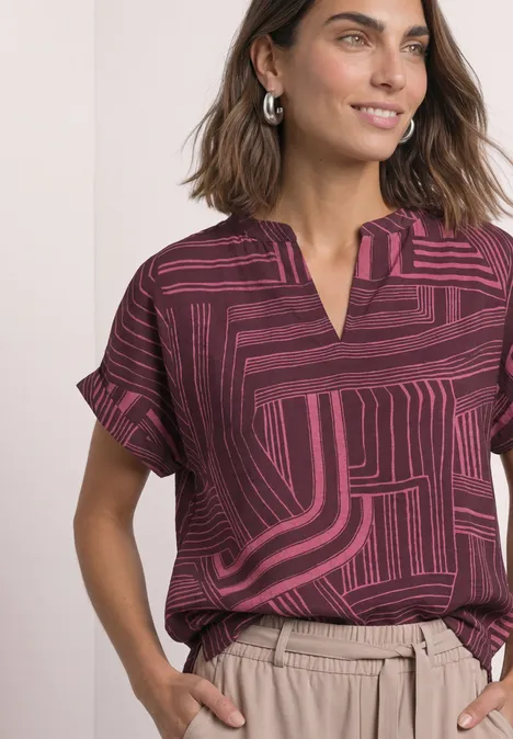Femme en blouse bordeaux à motifs géométriques, pantalon beige, boucles d'oreilles argentées, look décontracté.