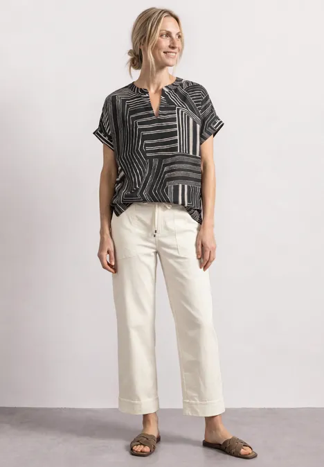 Frau trägt gemustertes schwarzes Shirt, helle Cropped-Hose und Sandalen, lächelnd stehend.