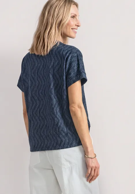 Vrouw van achteren in marineblauw shirt met golfmotief en lichte broek tegen neutrale achtergrond.