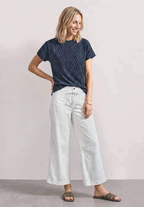 Vrouw staat voor neutrale achtergrond, draagt blauw gestreept T-shirt, witte wijde broek en sandalen.