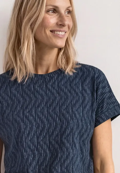 Bovenlichaam van een persoon met halflang blond haar en een wazig gezicht, gekleed in een donkerblauw shirt met golvende print