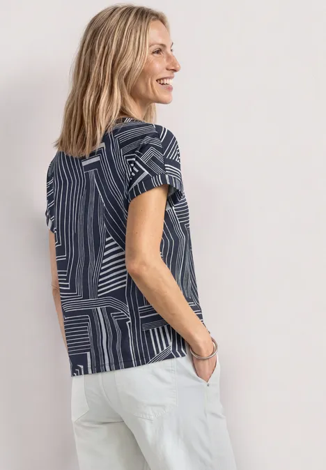 Vrouw in blauw patroon-t-shirt en witte broek, zijaanzicht tegen lichte achtergrond.