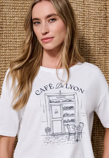 Personne porte un T-shirt blanc imprimé 'Café de Lyon' avec croquis de café devant un fond tressé