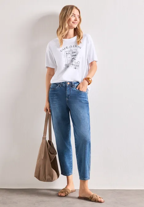 Femme en t-shirt blanc imprimé, jean bleu taille haute, sandales brunes et grand sac à main.