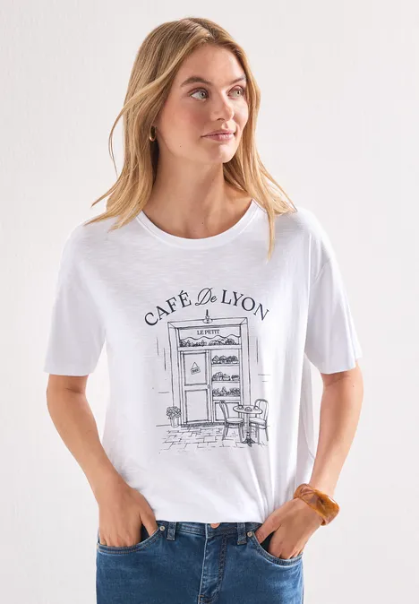 Femme en t-shirt blanc illustré « Café de Lyon », jean, cheveux lâchés, posant devant un mur clair.