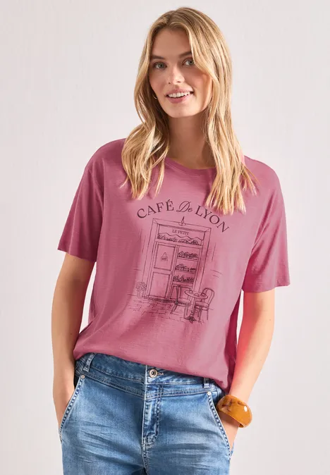 Femme blonde porte un t-shirt rose à motif café et un jean bleu, les mains dans les poches.