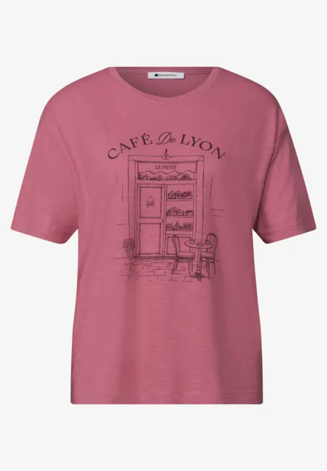 T-shirt rose orné d'un dessin d'entrée de café, deux chaises et l'inscription 'Café du Lion'.