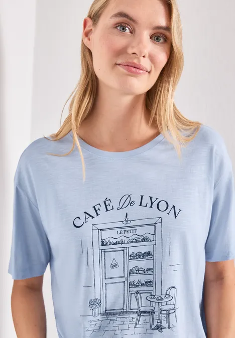 Femme porte un t-shirt bleu clair illustré 'Café de Lyon' avec table et chaise.