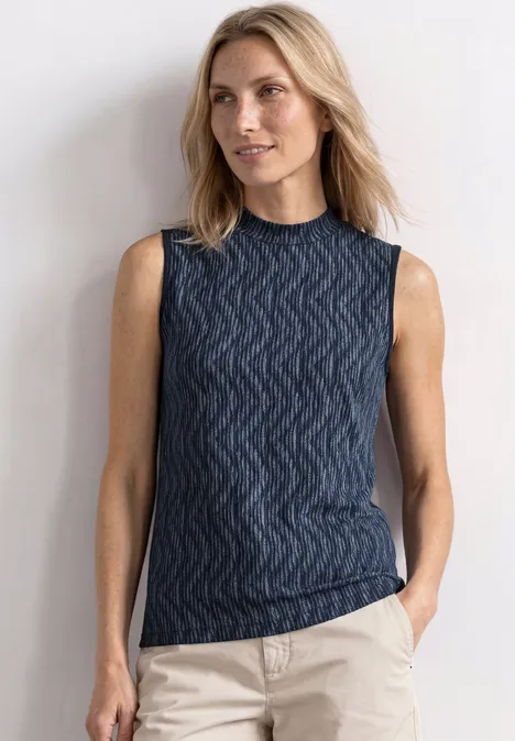 Top mit Turtle Neck und Print ashen blue Blonde Frau in ärmellosem, dunkelblauem, gemustertem Top und hellen Hosen vor hellem Hintergrund.