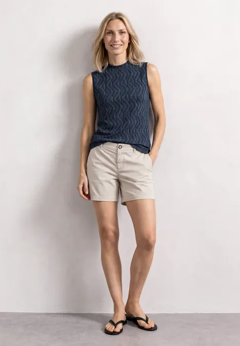 Top mit Turtle Neck und Print ashen blue Frau in blauem Top und beigen Shorts mit Flip-Flops, vor hellem Hintergrund, lächelt entspannt.