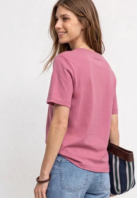 T-Shirt mit Rundhals und Chestprint blush berry Frau mit rosa T-Shirt und Jeans, lächelnd mit gestreifter Tasche in der Hand, steht vor hellem Hintergrund.