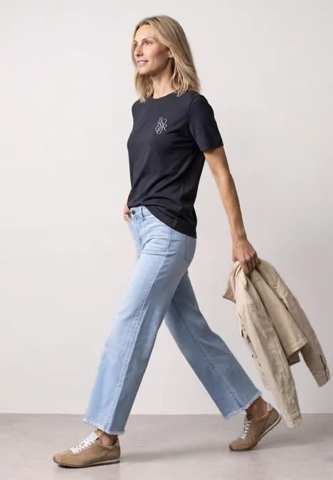Frau in dunkelblauem T-Shirt, hellblauen Jeans und Sneakers trägt beigen Mantel und geht vor weißem Hintergrund.