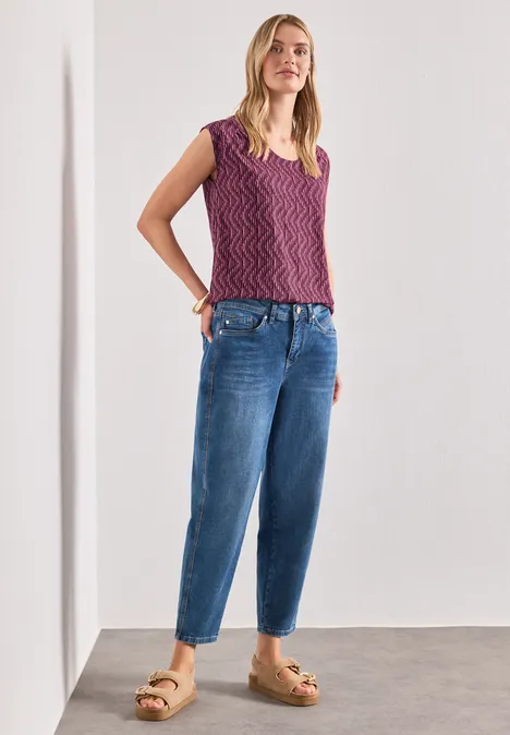 Shirt met afhangende schouders en elastische zoom blush berry Vrouw voor witte muur in lila gebreid topje, losse jeans en tan platformsandalen.