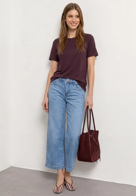 Basic Shirt met ronde hals in Unifarbe rustic rouge Vrouw leunt tegen witte muur, draagt paars shirt, wijde jeans, slippers en een rode tas.