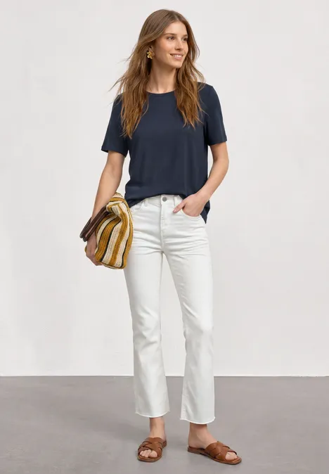 Basic T-Shirt mit Rundhals in Unifarbe darkest blue Frau in dunkelblauem T-Shirt, weißen Jeans, braunen Sandalen und gestreifter Tasche vor grauem Hintergrund.
