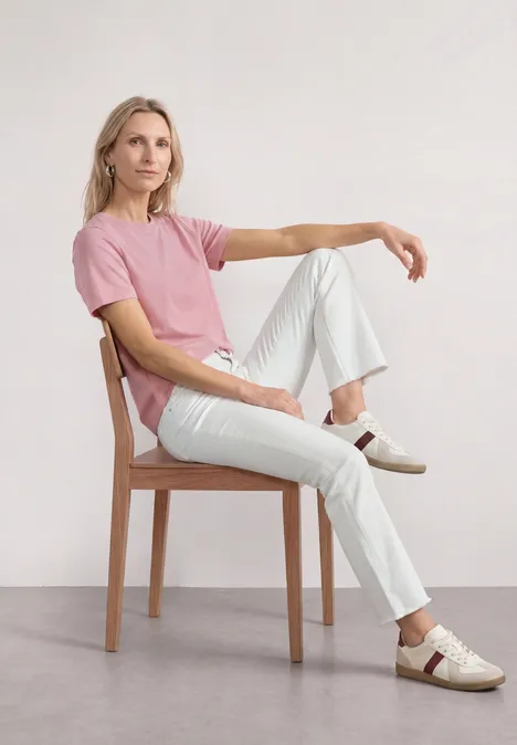 Basic T-Shirt mit Rundhals in Unifarbe vintage blush Frau in rosa T-Shirt und weißen Hosen sitzt seitlich auf Holzstuhl vor weißem Hintergrund.