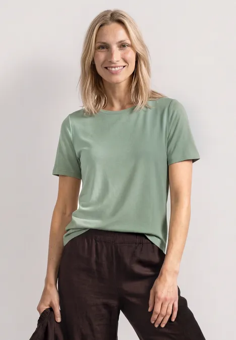 Basic Shirt met ronde hals in Unifarbe eucalyptus green Vrouw draagt een simpel mintgroen T-shirt en een bruine linnen broek tegen een lichte, rustige achtergrond.