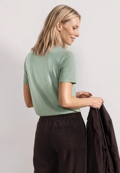 Basic Shirt met ronde hals in Unifarbe eucalyptus green Blonde vrouw van achteren in mintgroen t-shirt, die een zwarte jas vasthoudt.