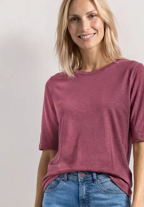 Basic T-Shirt mit Rundhals in Unifarbe dark clouded blush Lächelnde blonde Frau in kurzärmligem pinkem Shirt, das lässig in eine blaue Jeans gesteckt ist, vor hellem Hintergrund.