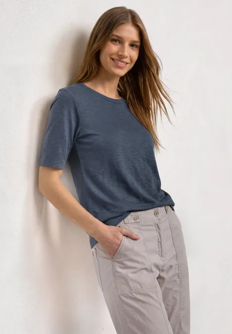 Frau lehnt an heller Wand, trägt dunkelblaues T-Shirt und hellbeige Hose, eine Hand in der Tasche.
