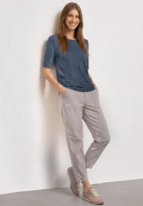 Frau lehnt lässig an einer Wand, trägt blaues T-Shirt, hellgraue Hose und beigefarbene Sneaker.