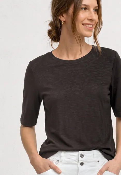 Frau trägt dunkelgraues T-Shirt und weiße Hose mit Knöpfen, Hände in den Taschen.