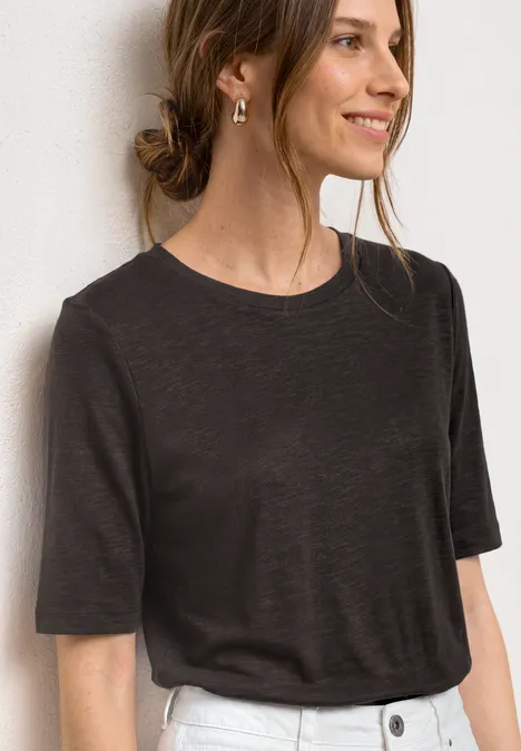 Frau in dunkelgrauem T-Shirt mit goldenem Ohrring und Halskette vor weißem Hintergrund